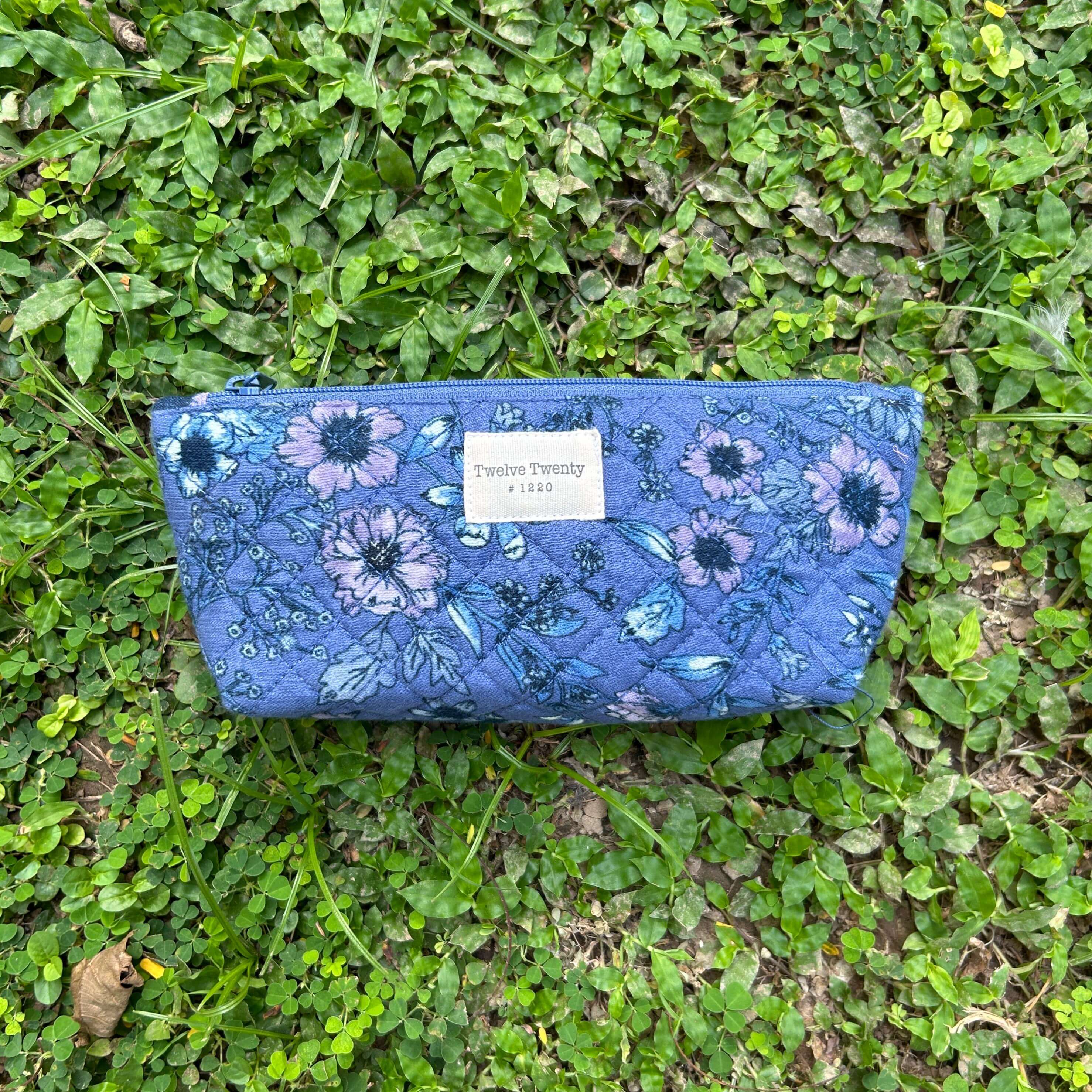 Blue floral makeup pouch with zipper, featuring a quilted design, lying on green grass.