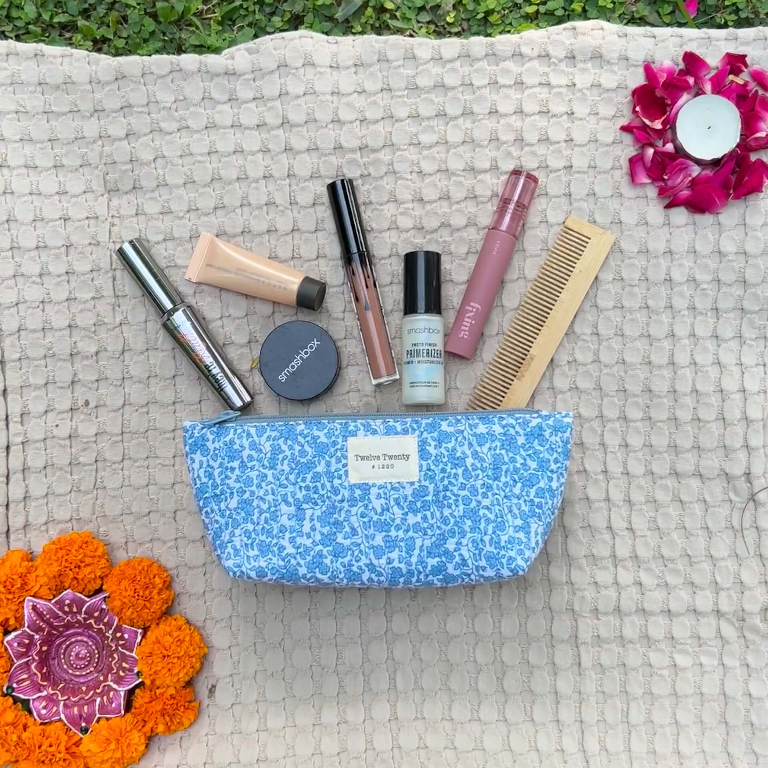 Blue makeup pouch with makeup products and comb on textured mat, surrounded by flowers, highlighting cosmetic bag versatility.