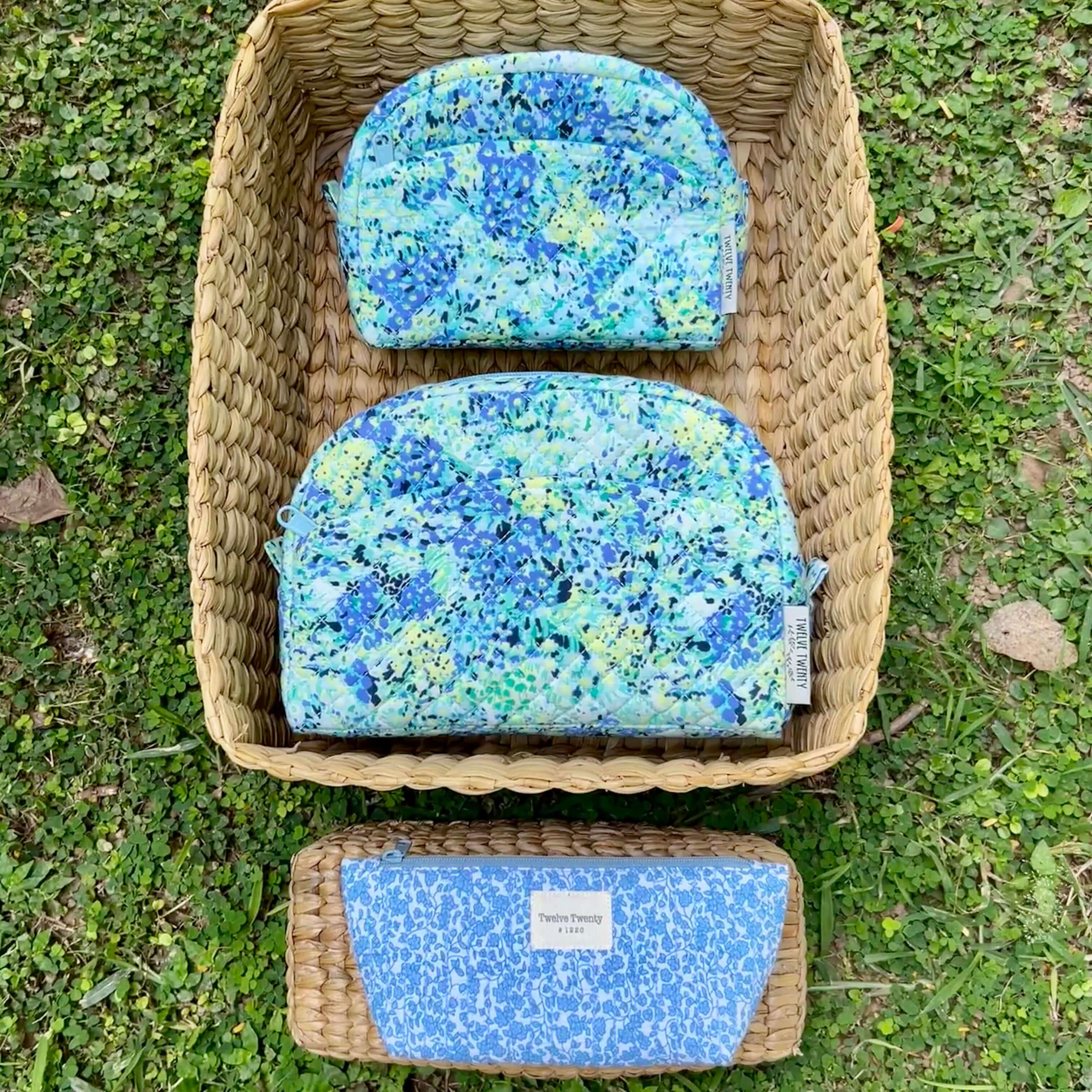 Trinity set with two blue floral makeup bags and a blue everyday pouch in a wicker basket on grass. Perfect cosmetic storage solution.